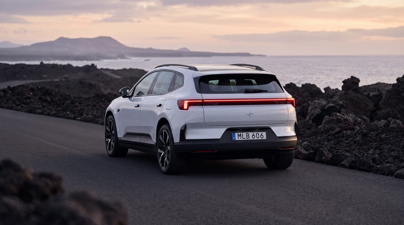 SUV electric Polestar 4 alb, în trei sferturi spate, pe un drum de coastă, cu luneta noii versiuni vizibilă
